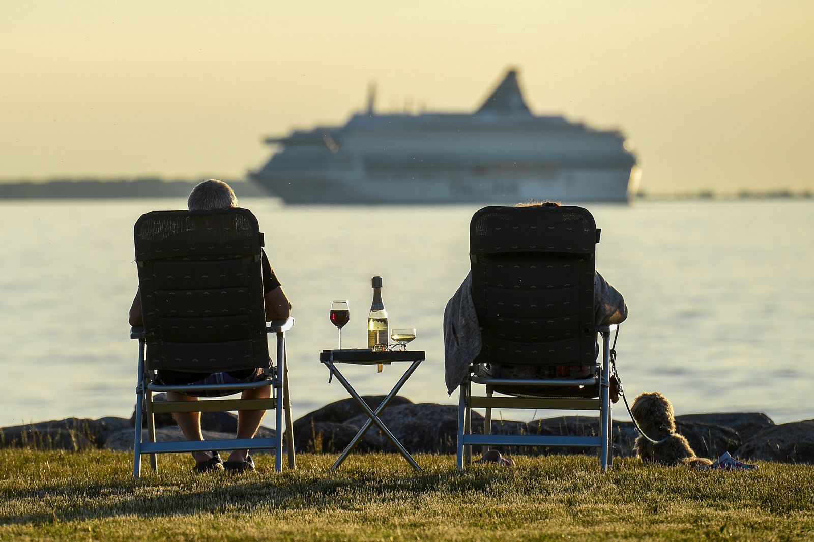 Two chairs at the water&rsquo;s edge facing a passing cruise ship