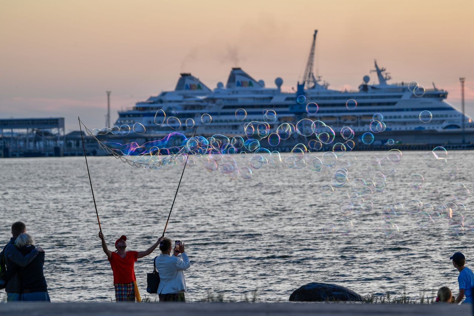 People making giant soap bubbles on the shore as a cruise ship sits in the harbor